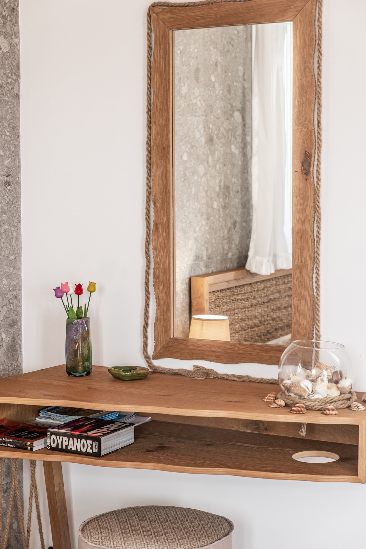 A wooden console table is positioned against a wall, featuring a large, natural-finish mirror with rope detailing. A small vase holds fresh flowers, while a bowl contains shells. Travel magazines rest neatly on the shelf, accompanied by a decorative dish.