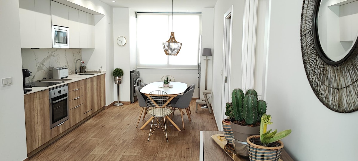 An open kitchen and dining area is featured, designed with modern cabinetry and a sleek countertop. A round dining table is surrounded by stylish chairs, complemented by potted plants. Natural light enters through a large window, illuminating the neutral-colored space.