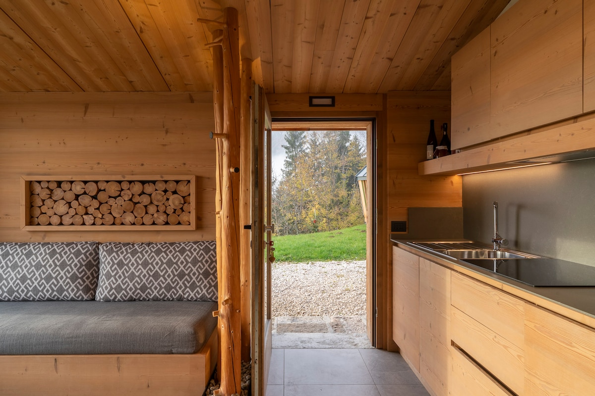 A view from inside the lodge focuses on the wooden interior, featuring a cozy seating area with a textured gray cushion. The kitchen area is equipped with modern cabinets and a stainless steel sink. Natural light enters through the open door, highlighting the outdoor greenery.