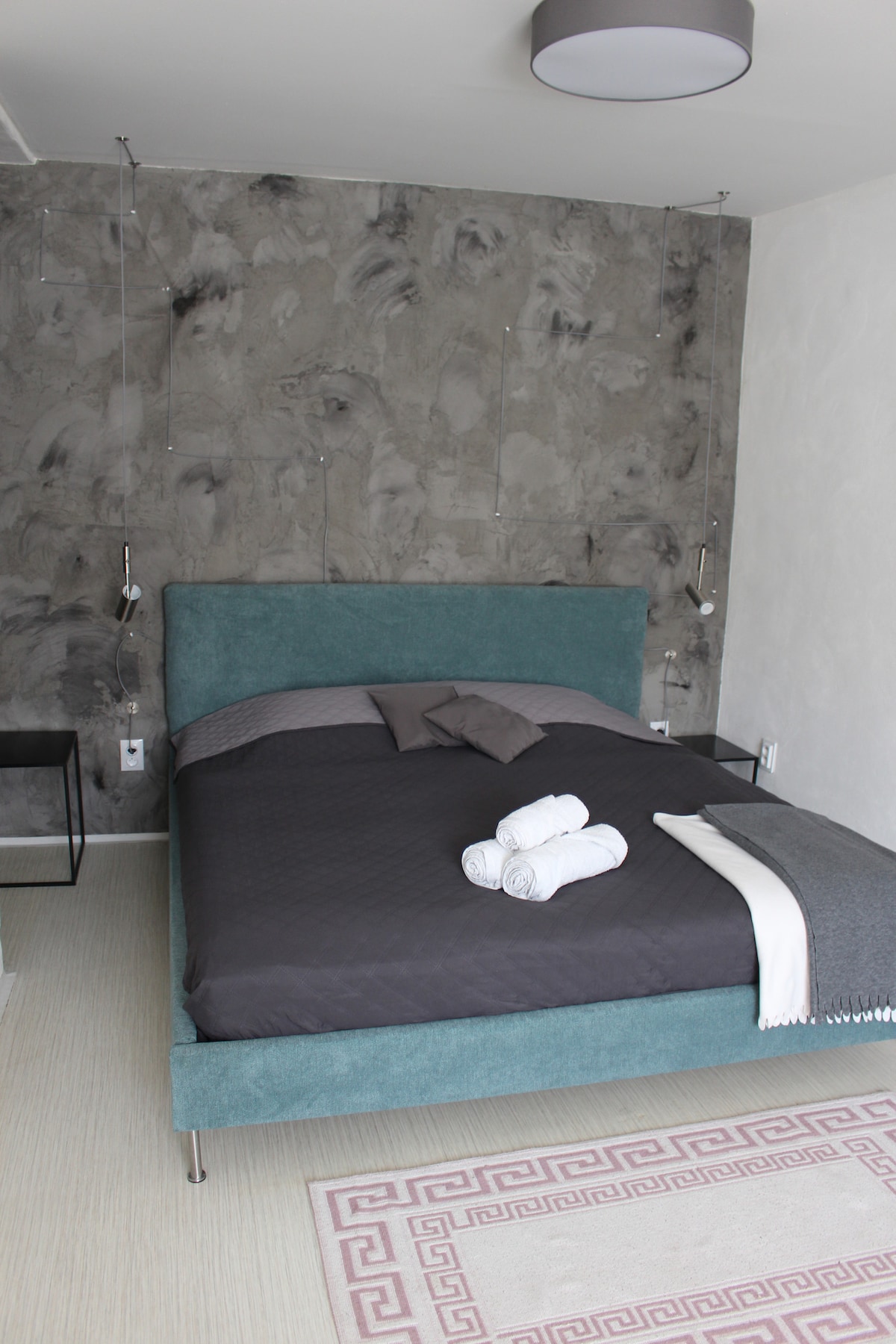 A spacious bedroom features a modern grey wall and a plush bed dressed in dark linens. Neatly arranged towels are placed on the bed. A small bedside table sits beside the bed, providing a simple functional touch to the room.