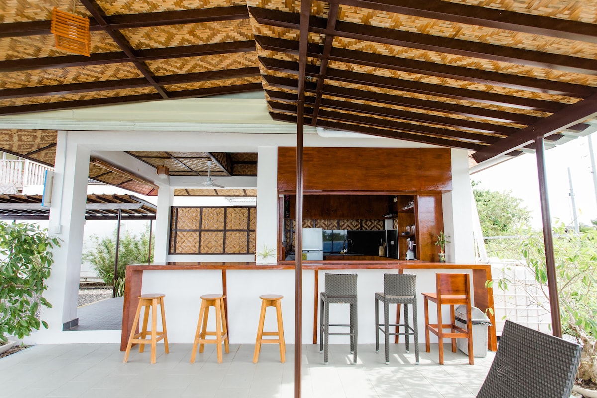 A spacious outdoor dining area features a wooden bar with accompanying stools, set beneath a thatched roof. The open design allows for natural light and fresh air, creating a welcoming space for guests to relax and enjoy their surroundings.