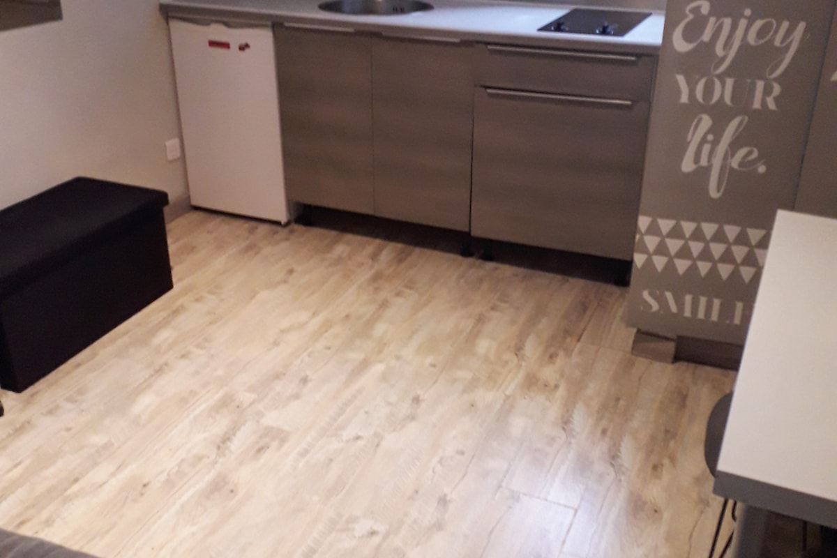 A functional kitchenette features modern cabinetry with a sink and a stove. A compact refrigerator is visible alongside a black storage bench. The flooring is wooden, contributing to a clean and contemporary look. A light wall with motivational text adds subtle character.