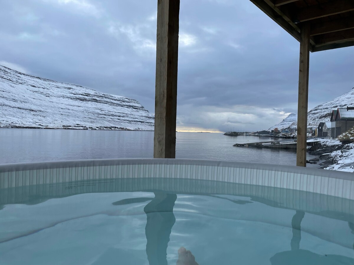 An outdoor spa is situated on a wooden deck, offering views of serene water and snow-covered hills. The water appears calm, reflecting cloudy skies and distant land, creating a tranquil atmosphere for relaxation.