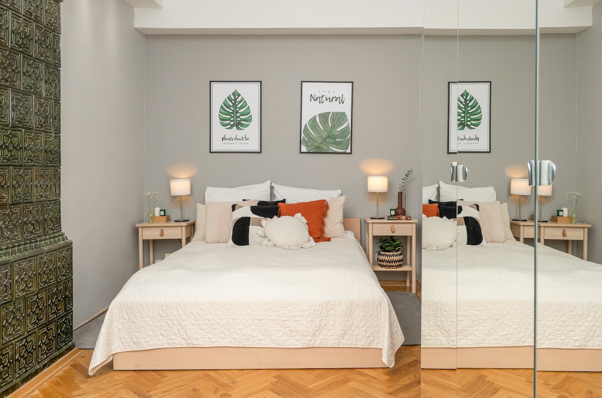 A spacious bedroom is depicted, featuring a light beige quilted bedspread and soft pillows in various colors. Flanking the bed are two nightstands with lamps and decorative plants. The room is enhanced by framed green leaf prints on the wall, and a mirrored wardrobe reflects the calming space.