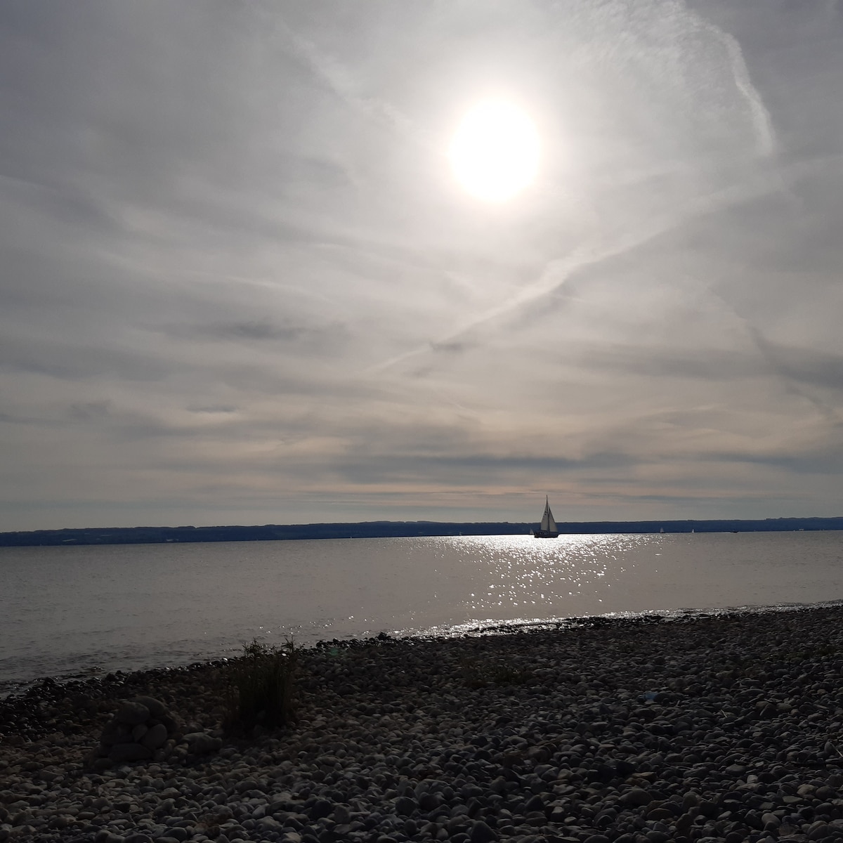 A sunlit lake scene features a gentle reflection shimmering on the water's surface. A sailboat glides smoothly across the horizon, with rocky shorelines and distant land visible under a cloudy sky.