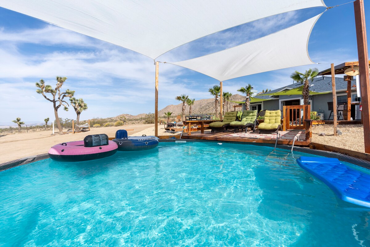A refreshing swimming pool is surrounded by a sunshade, with pool floats in pink and blue. Comfortable lounge chairs are positioned beside the pool, offering a space for relaxation. The view extends to the desert landscape, punctuated by palm trees and distant mountains.