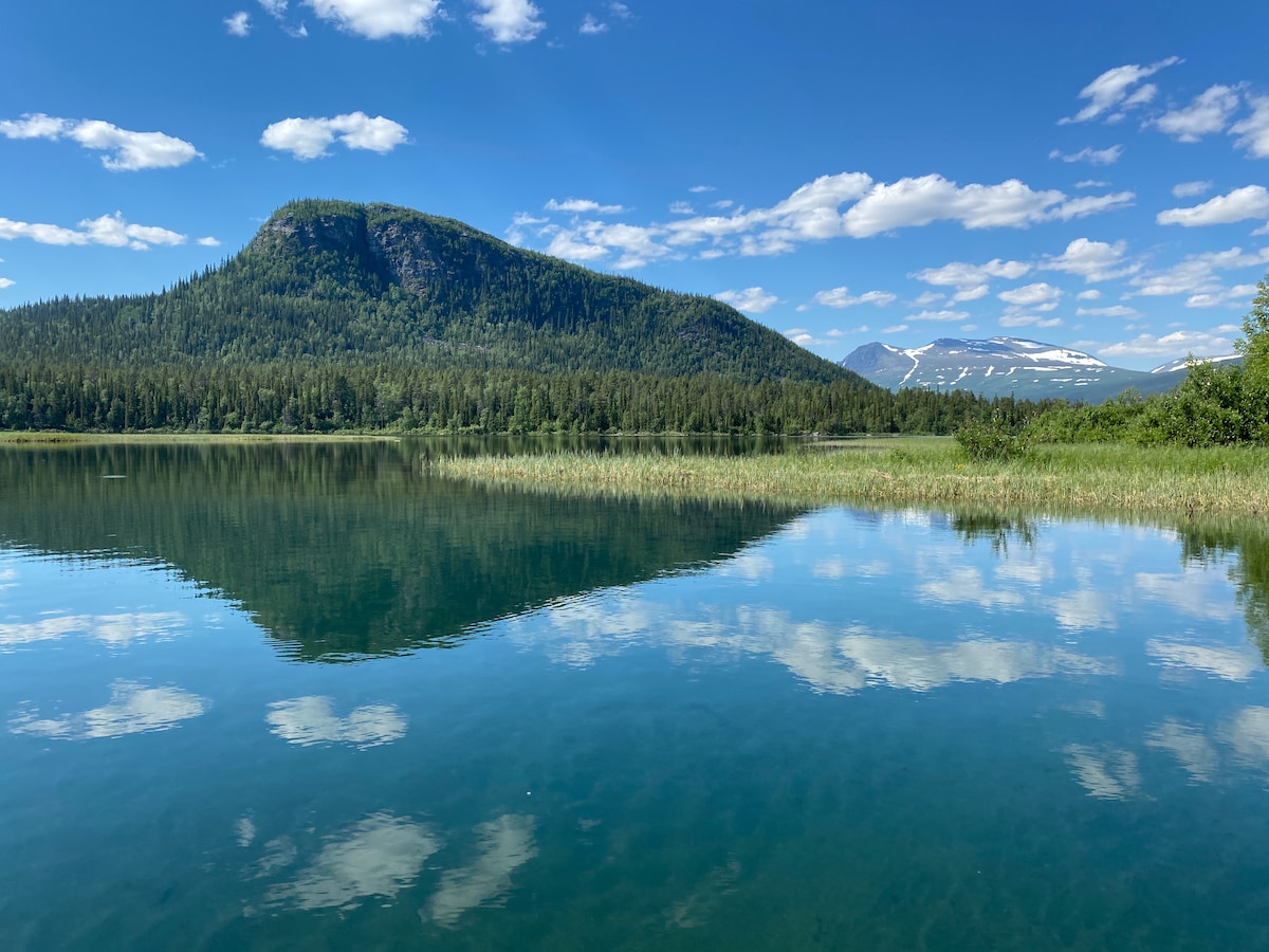 A serene lake is captured reflecting a lush green mountain and clear blue sky with scattered clouds. The calm water mirrors the surrounding forest and distant peaks, creating a peaceful landscape that highlights the natural beauty of the area.