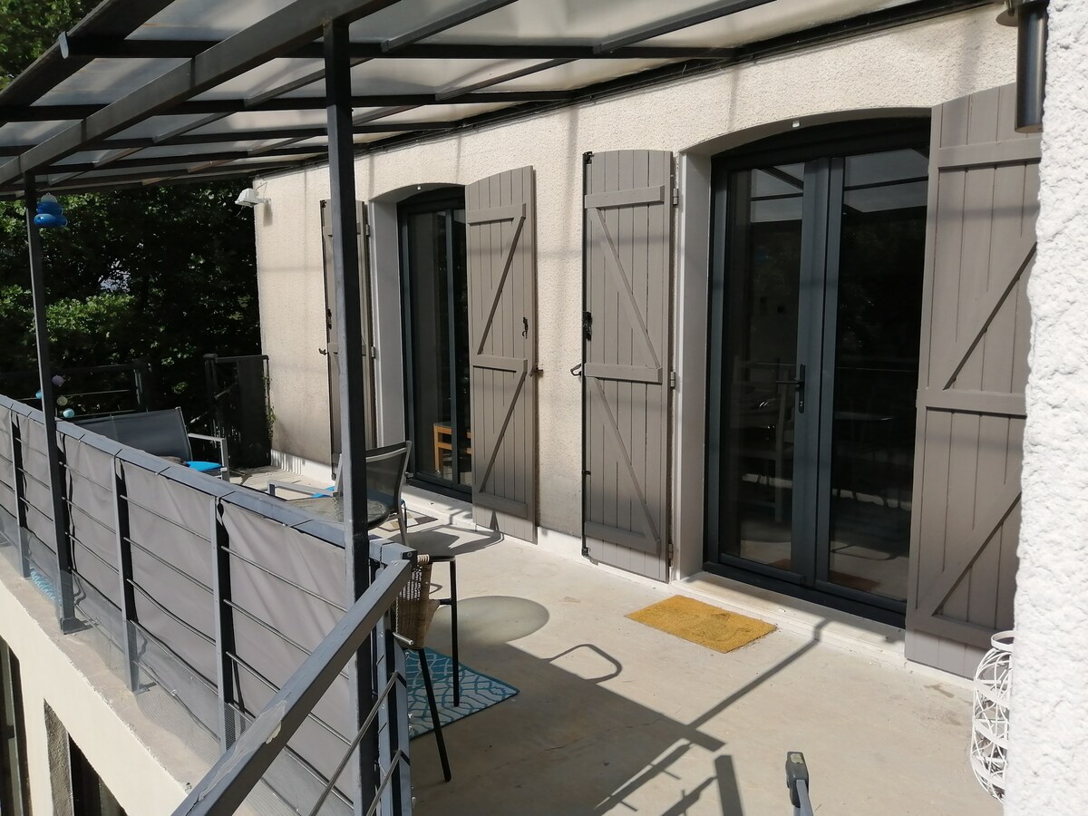A private terrace features a covered area with outdoor seating. Double doors provide access to the apartment, framed by wooden shutters. The terrace is partially shaded, with a textured mat and a small decorative rug visible on the ground.