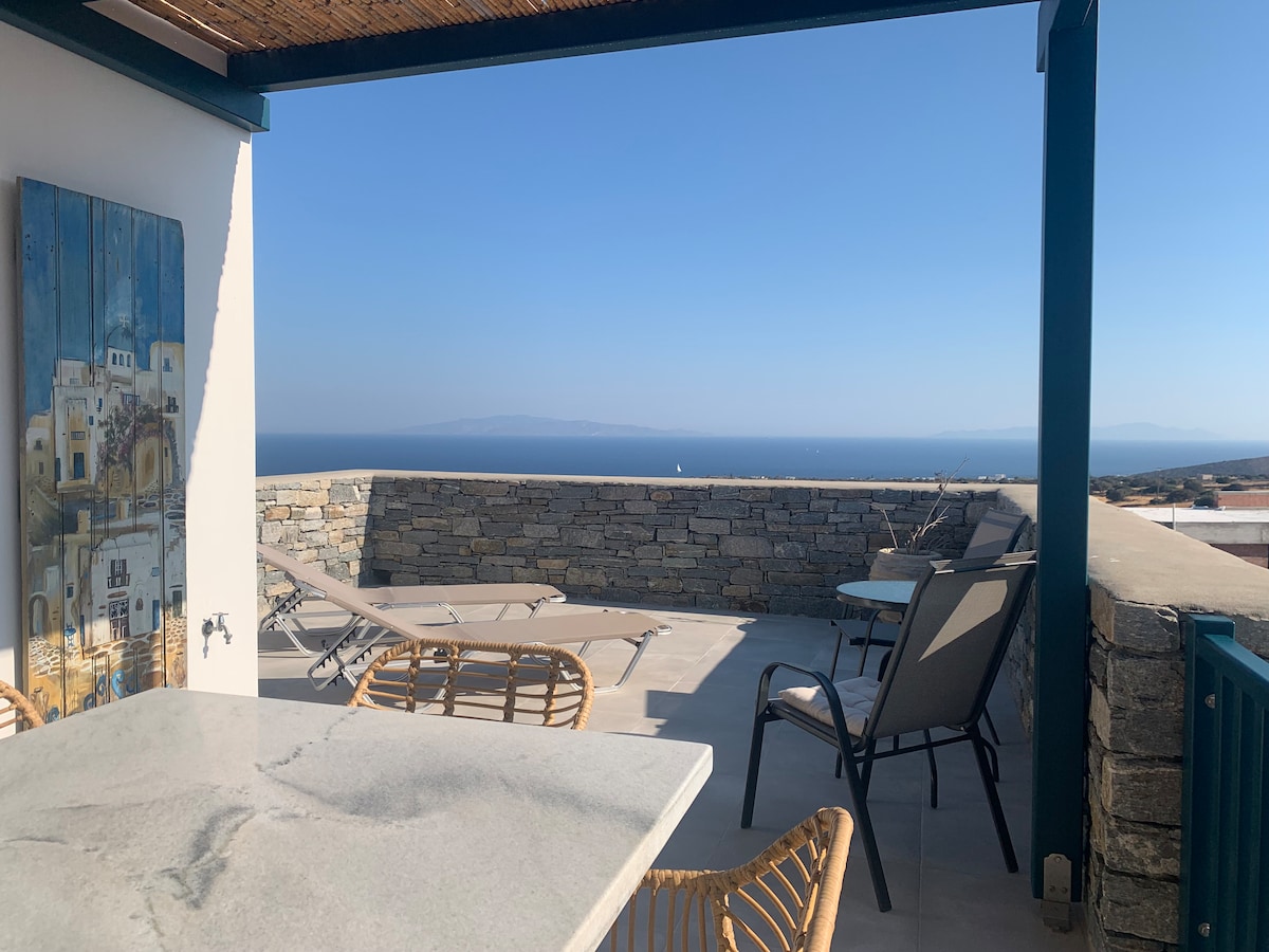 A spacious terrace is shown, featuring a table and several chairs. Sun loungers are positioned nearby, with stunning views of the blue sea and distant islands in the background. The stone railing complements the open sky, offering a serene atmosphere.
