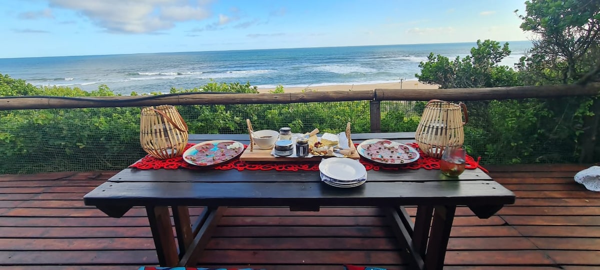 A wooden dining table is set with plates, glasses, and a serving tray, overlooking the ocean. Two lanterns provide soft light, while a vibrant table runner adds a touch of color. The view includes gentle waves and lush greenery in the background.