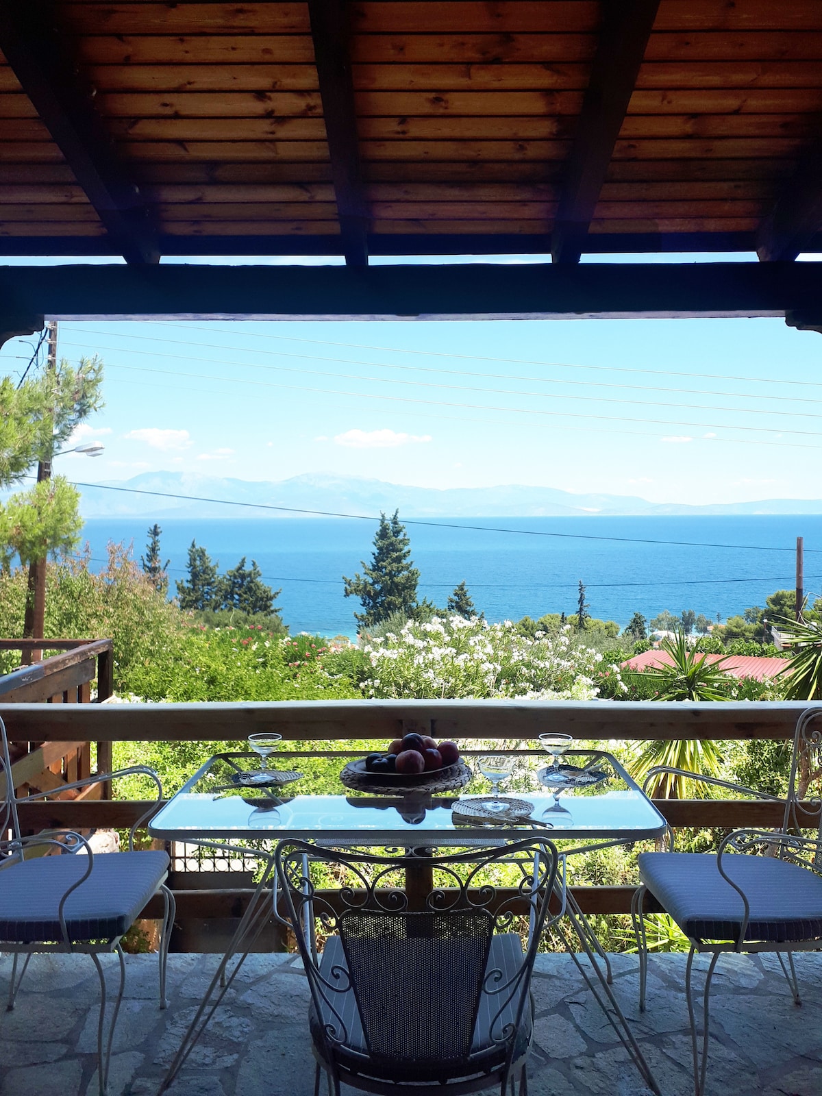 A dining table is set on a covered terrace, showcasing seating for four. The table is adorned with a fruit bowl. In the background, a panoramic view of the sea and surrounding greenery is visible, framed by wooden beams.