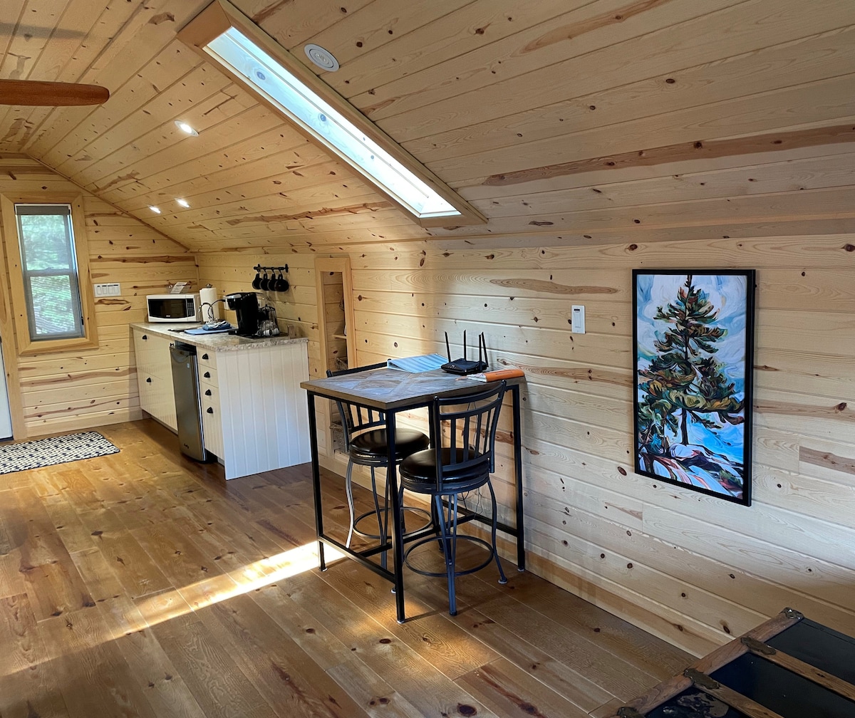 An open concept area features wood-paneled walls and a cozy sitting space with two black chairs at a round table. A kitchenette with a microwave and coffee station is seen against one wall. Natural light filters through a skylight, enhancing the inviting ambiance.