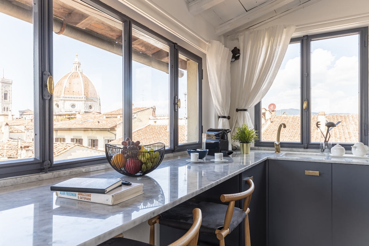 A bright kitchen area is displayed with large windows overlooking the rooftops of Florence and the Duomo. The countertop features a bowl of fruit, coffee machine, and stoneware, enhancing the functional and stylish layout.