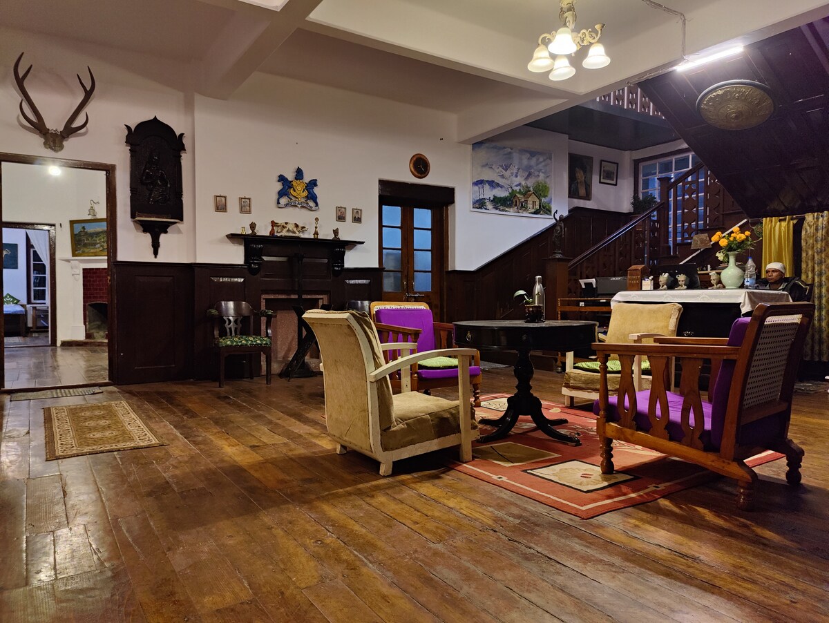 A spacious living area features wooden flooring and a variety of seating options, including chairs with colorful cushions and a low coffee table. Wall art and decorative elements provide character, while an elegant staircase is visible in the background. The room is softly illuminated by overhead lighting.