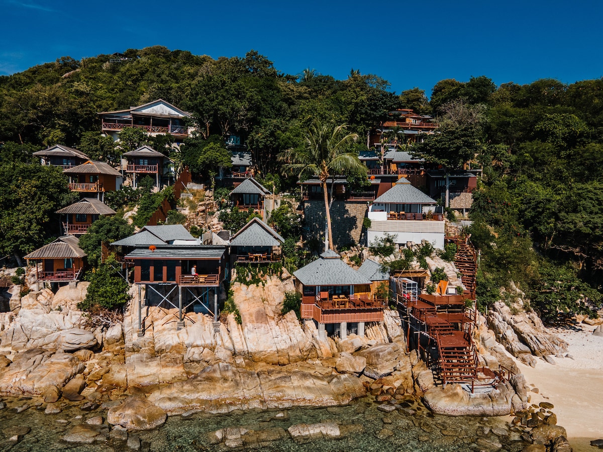 A collection of beachside villas is nestled into a rocky coastline, surrounded by lush greenery. Each villa features a distinct roof design, with wooden decks overlooking the water, enhancing the coastal scenery.