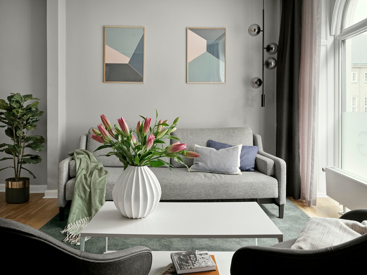 A contemporary living room features a gray sofa adorned with decorative pillows. A white coffee table is positioned in the center, complemented by a vase filled with fresh flowers. Two modern artworks hang on the wall, while a leafy plant adds a touch of greenery.