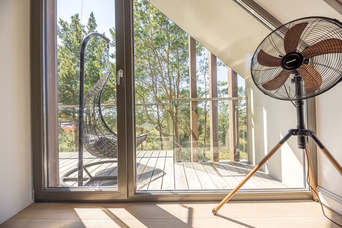 A well-lit indoor space features large glass doors opening to a wooden deck. A modern hanging chair is placed on the deck, surrounded by greenery. An industrial-style fan stands nearby, adding a functional element to the scene.