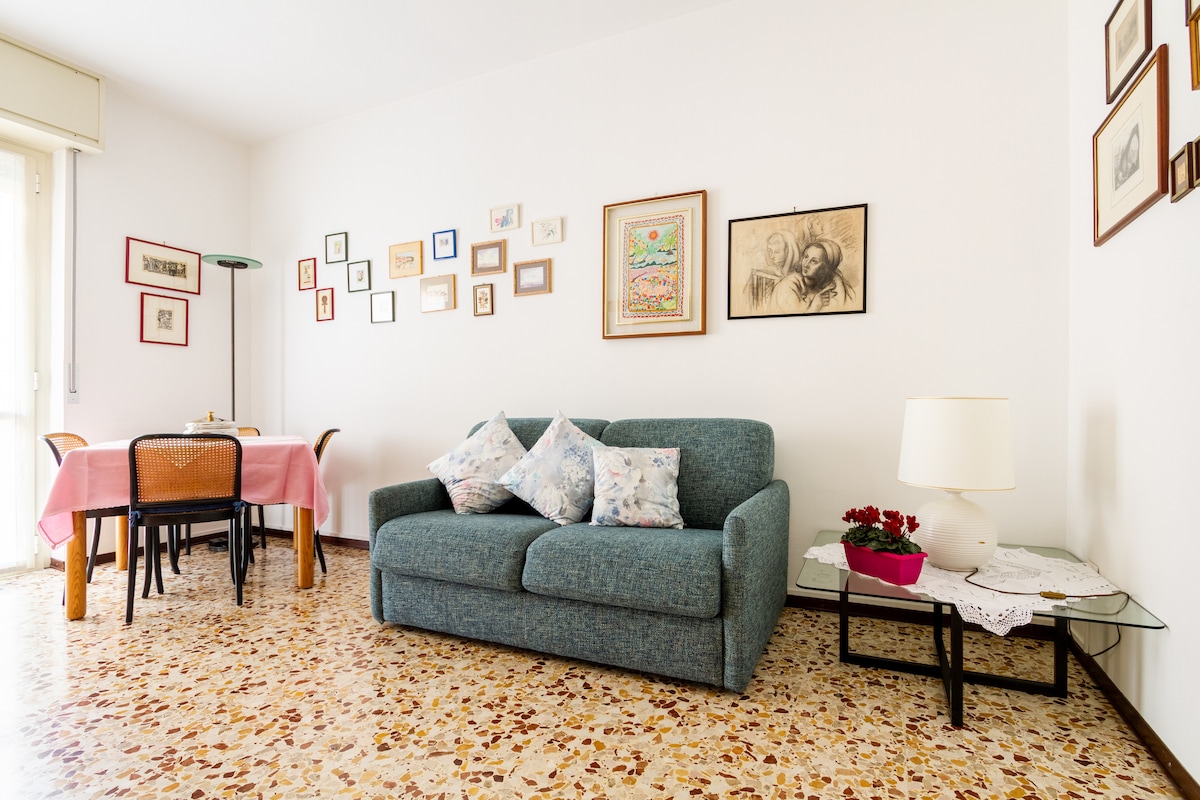 A living area is showcased with a comfortable sofa adorned with decorative pillows. A simple dining table is set nearby, complemented by wooden chairs. The walls are decorated with a variety of framed artworks, and a plant sits on a side table, creating a cozy atmosphere.