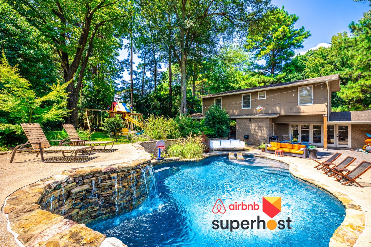 The backyard features a serene salt-water pool with a cascading waterfall, surrounded by lounge chairs. Lush greenery provides privacy, while a vibrant playground is visible in the background. The modern home overlooks this inviting outdoor space, offering a tranquil retreat.