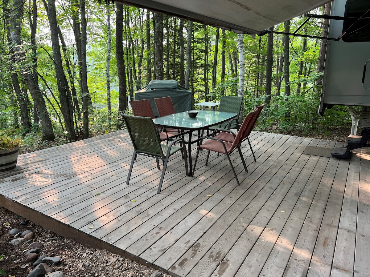 A wooden deck is surrounded by lush green trees, featuring a glass-top table and six chairs. A grill is positioned in the background, and the natural scenery enhances the outdoor dining space.