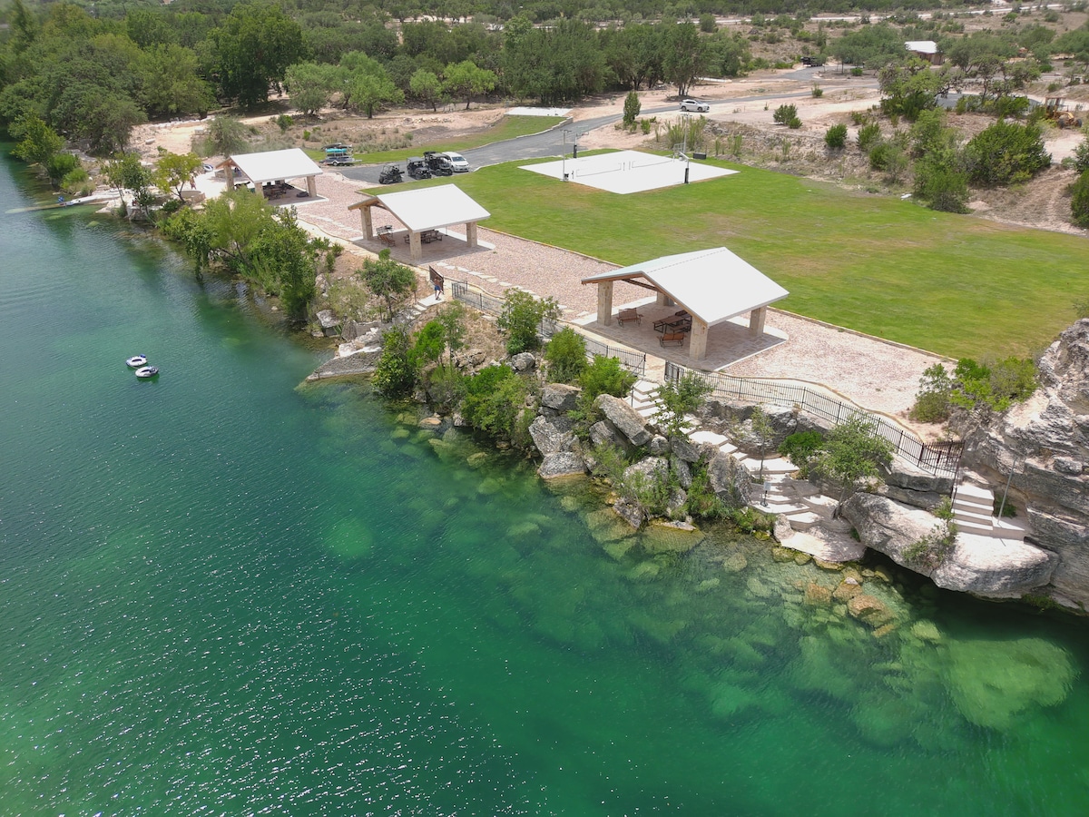 Aerial view of the riverfront area showing private pavilions on manicured grass, surrounded by trees. A clear section of the Nueces River displays visible rocks and aquatic plants. Further back, basketball and pickleball courts are located near parking space.