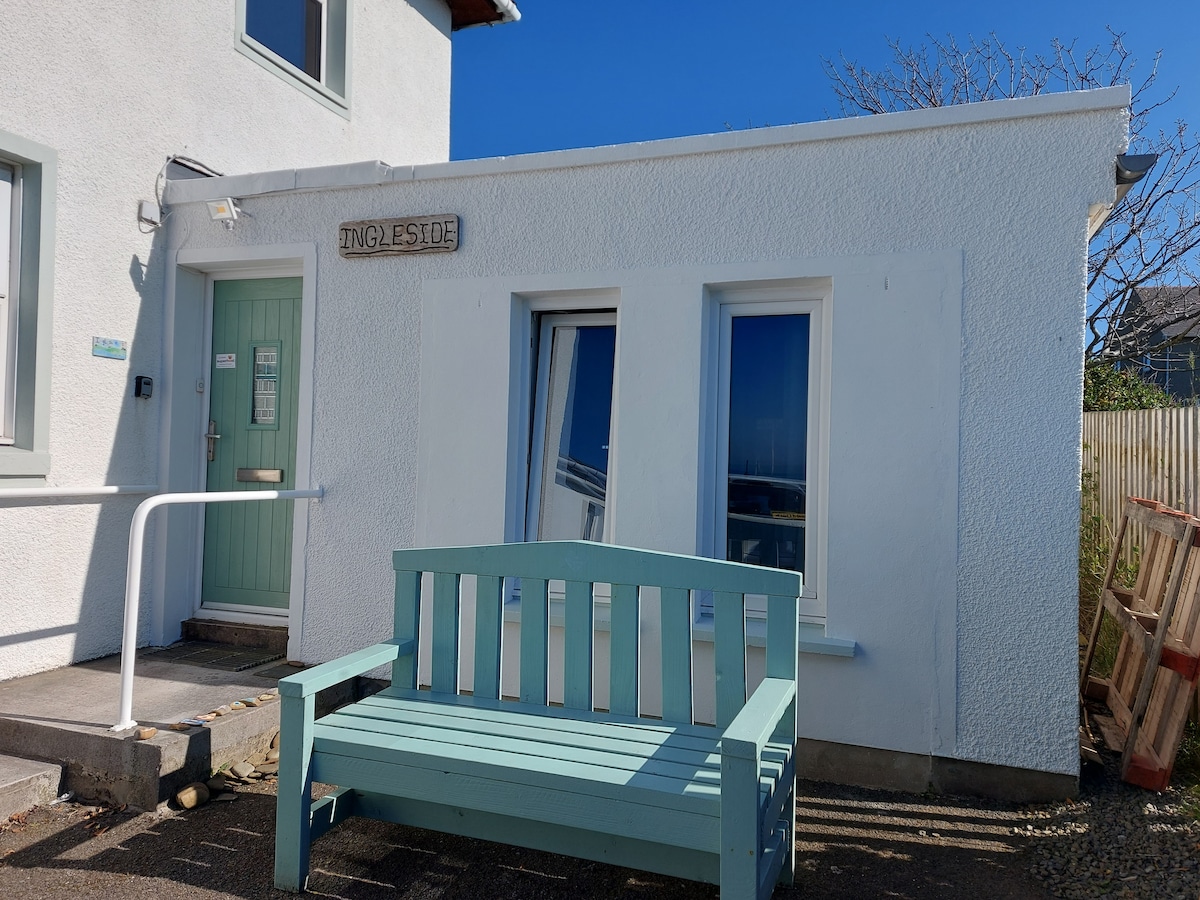 The exterior of the studio apartment displays a light-colored façade with a welcoming blue bench positioned in front. Large windows reflect the surrounding scenery, allowing natural light to enter the space. A leafy area is visible beside the entrance.