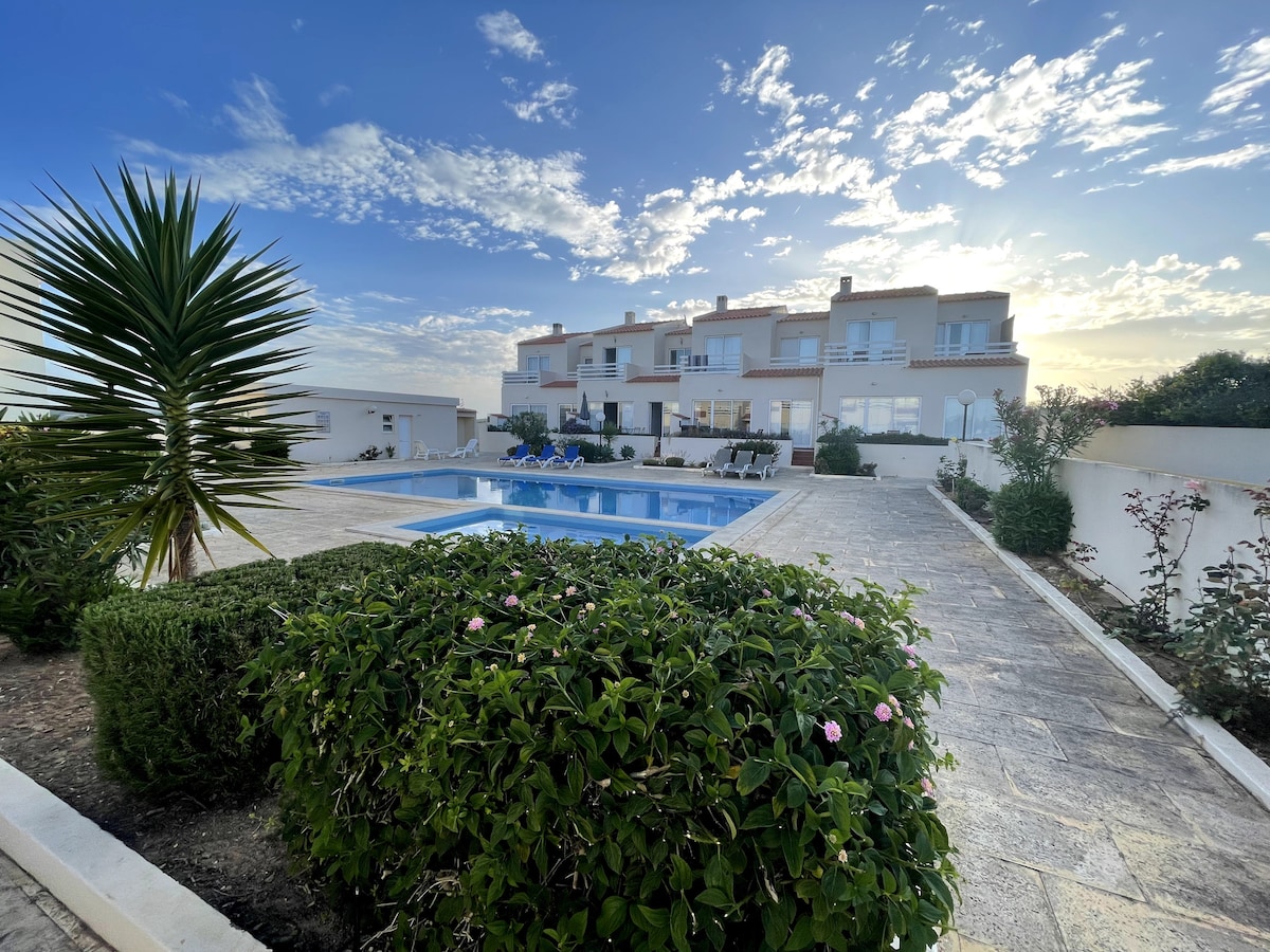 The image captures a tranquil villa surrounded by lush greenery and a beautifully maintained pool area. The expansive terrace features lounge chairs and tables, ideal for relaxation. The sky displays soft hues as the sun sets, enhancing the peaceful ambiance of the outdoor space.