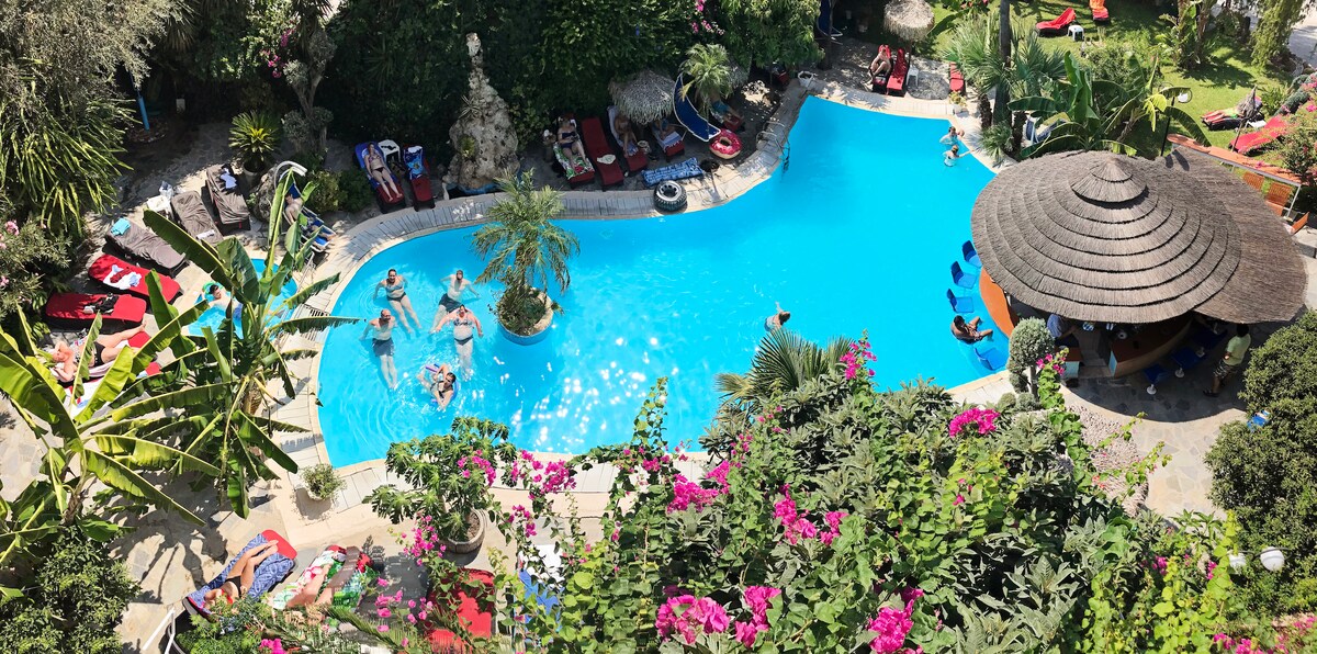 A serene swimming pool is surrounded by lush greenery and vibrant flowers. Sun loungers in red and blue hues are placed around the pool. A thatched-roof structure offers shade nearby. Guests enjoy the water, enhancing the tranquil environment of the property.