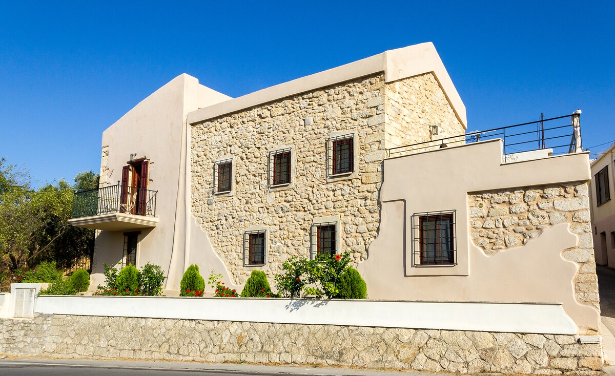 The exterior of a charming stone mansion is displayed, featuring textured stone walls and a light-colored façade. A balcony is visible above the entrance, framed by dark wooden shutters. Neat landscaping with shrubs and flowers enhances the inviting appearance of the property.