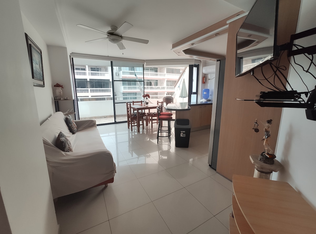 A bright living area is presented with a large window extending across one wall, providing ample natural light. A comfortable sofa bed is positioned beside a dining table set for four. The space features modern furnishings and a clean, tiled floor, enhancing the contemporary feel.