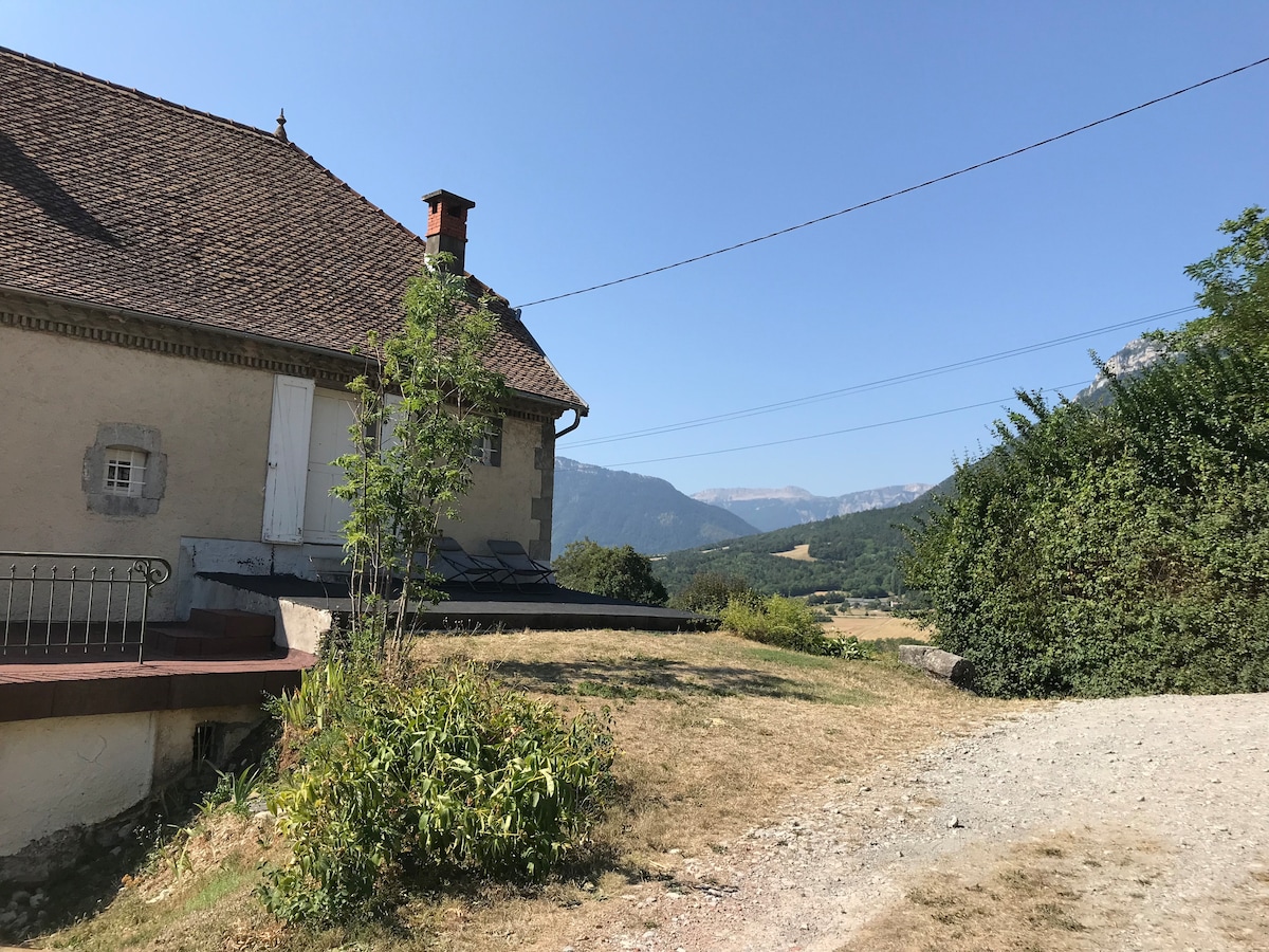 A side view of a quaint house features a sloped roof and a stone facade, framed by surrounding greenery and a gravel pathway. The distant mountains rise in the background under a clear blue sky, offering a serene rural landscape.