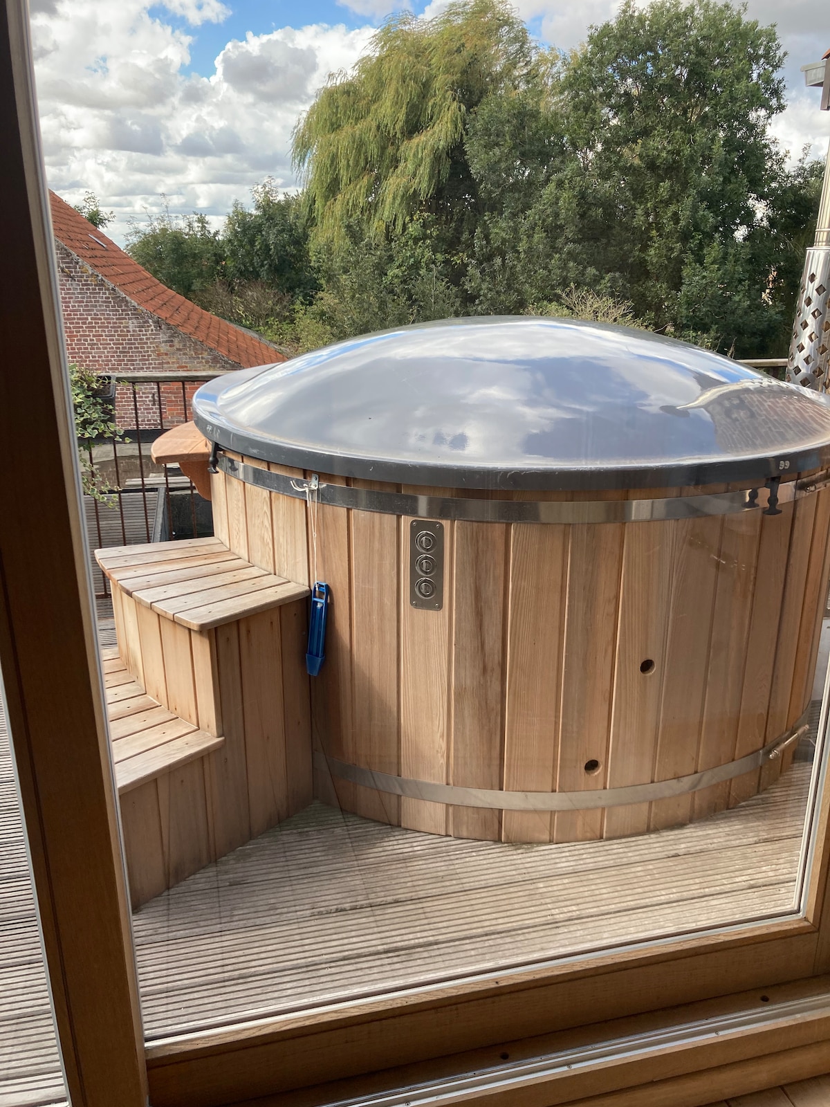 A wooden hot tub with a clear domed cover is positioned on a wooden deck. Steps lead up to the tub, providing easy access. The surrounding greenery and trees are visible, creating a calm outdoor atmosphere.