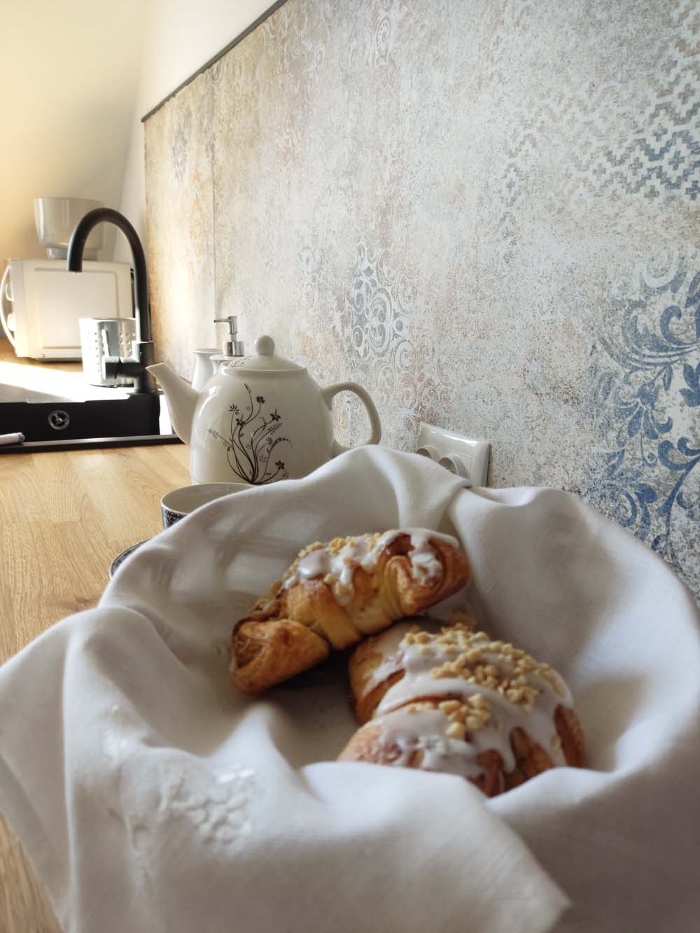 A woven basket lined with a white cloth holds two freshly baked croissants, one drizzled with icing and topped with crushed nuts. A teapot and cup are positioned on a wooden countertop, complemented by a decorative tiled wall in soft colors.