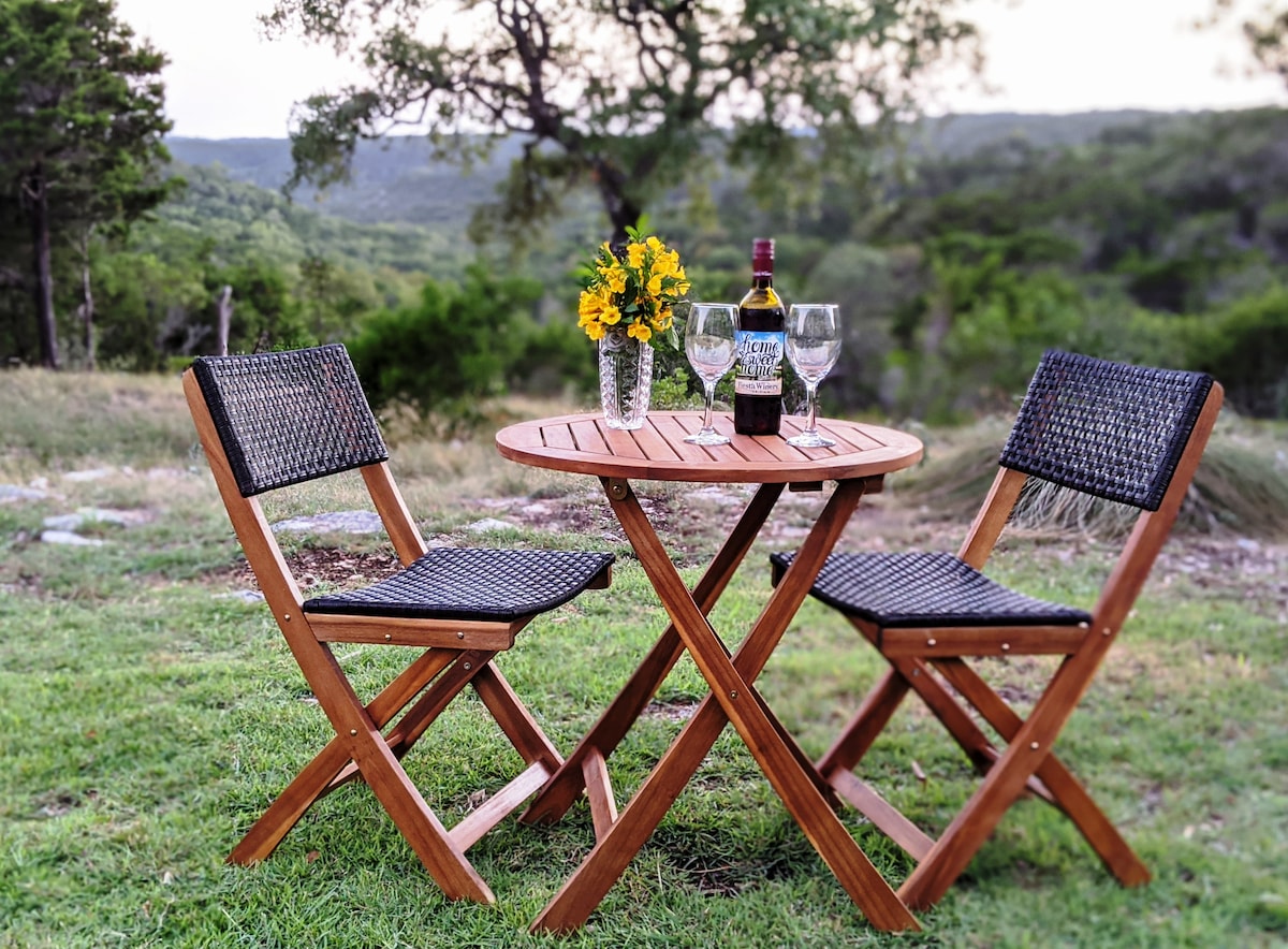 An outdoor seating arrangement features a small wooden table surrounded by two folding chairs. A vase with bright flowers adds color to the table, which also holds a bottle of wine and two glasses. The lush green landscape and distant hills create a serene backdrop.