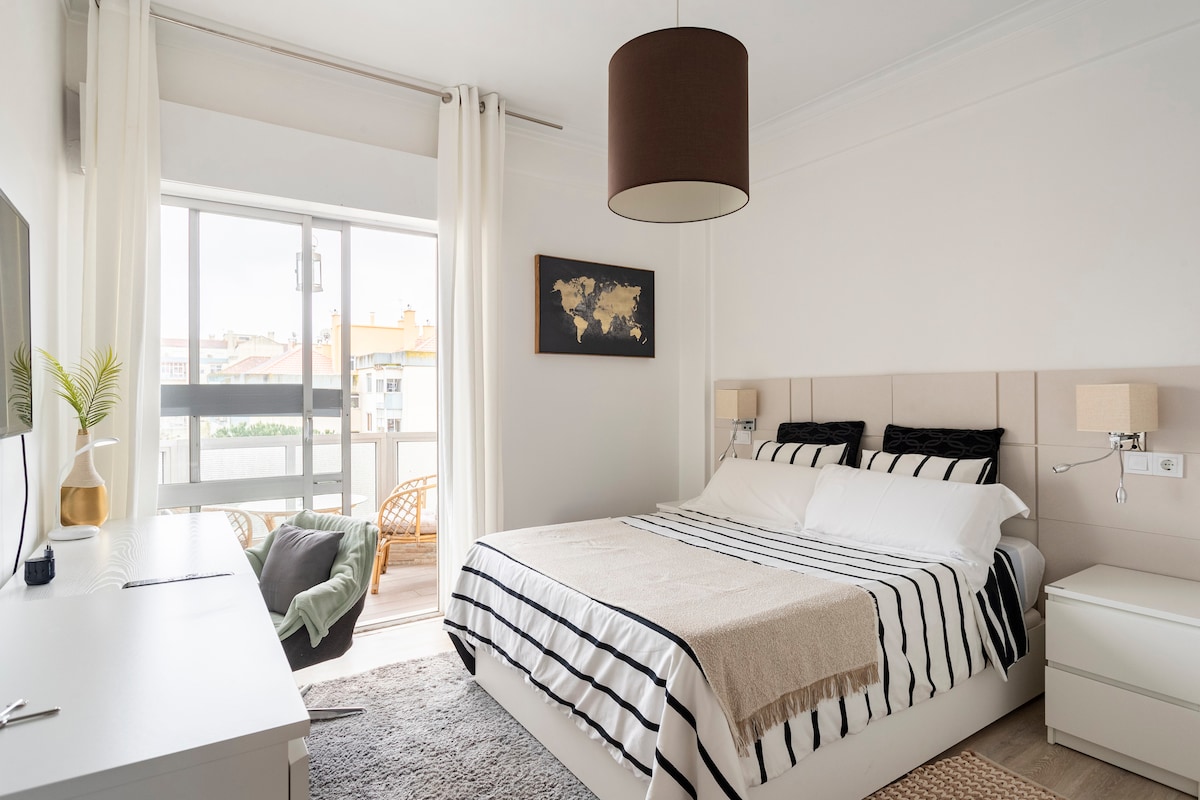 A bright room features a spacious bed with a striped duvet and pillows. Natural light enters through large windows, complemented by sheer curtains. A wooden chair and a small table are positioned near the window. A light-colored dresser adds functional storage.