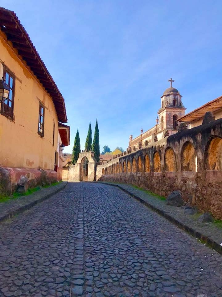 A cobblestone street is bordered by historic buildings, featuring a traditional adobe structure on the left and a church with a prominent bell tower on the right. Lush, tall fir trees stand in the background, contributing to the serene atmosphere.