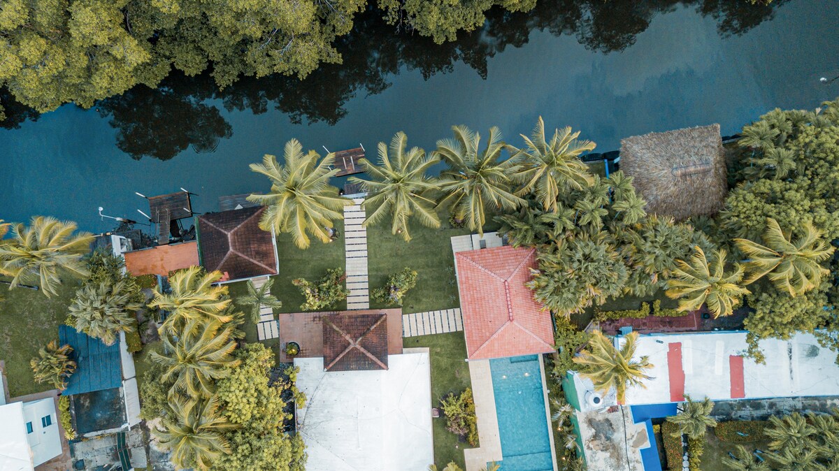 An aerial view captures a tranquil property along a canal, surrounded by lush greenery and palm trees. The spacious garden features a private pool and a pathway leading towards the water's edge, enhancing the natural beauty of the setting.