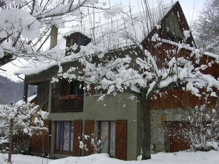 A charming duplex house is surrounded by a snowy landscape, showcasing a mix of stone and wood architecture. Snow-covered trees frame the building, with a balcony on the upper level and wooden shutters adorning the windows. The cold, quiet scene evokes a sense of winter tranquility.