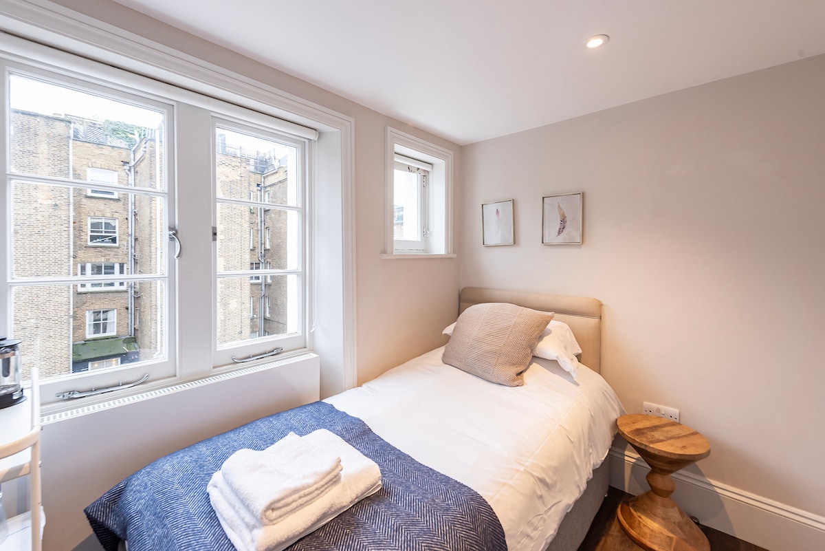 A compact sleeping area features a spacious bed with a light-colored duvet and a textured navy blanket. Two windows provide natural light and views of neighboring buildings. A small wooden stool accompanies the bed, and three framed artwork pieces adorn the wall.