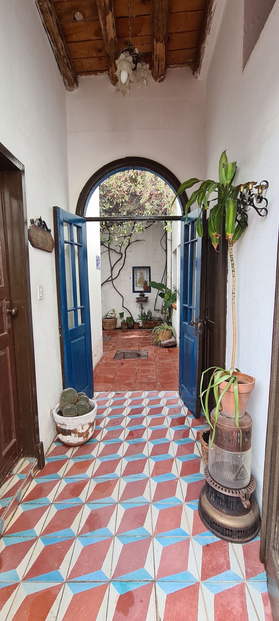 A bright entryway features double blue doors that open to a quaint courtyard filled with greenery. The floor is adorned with colorful patterned tiles, complementing the rustic wooden ceiling. Potted plants and decorative elements add charm to this inviting passage.