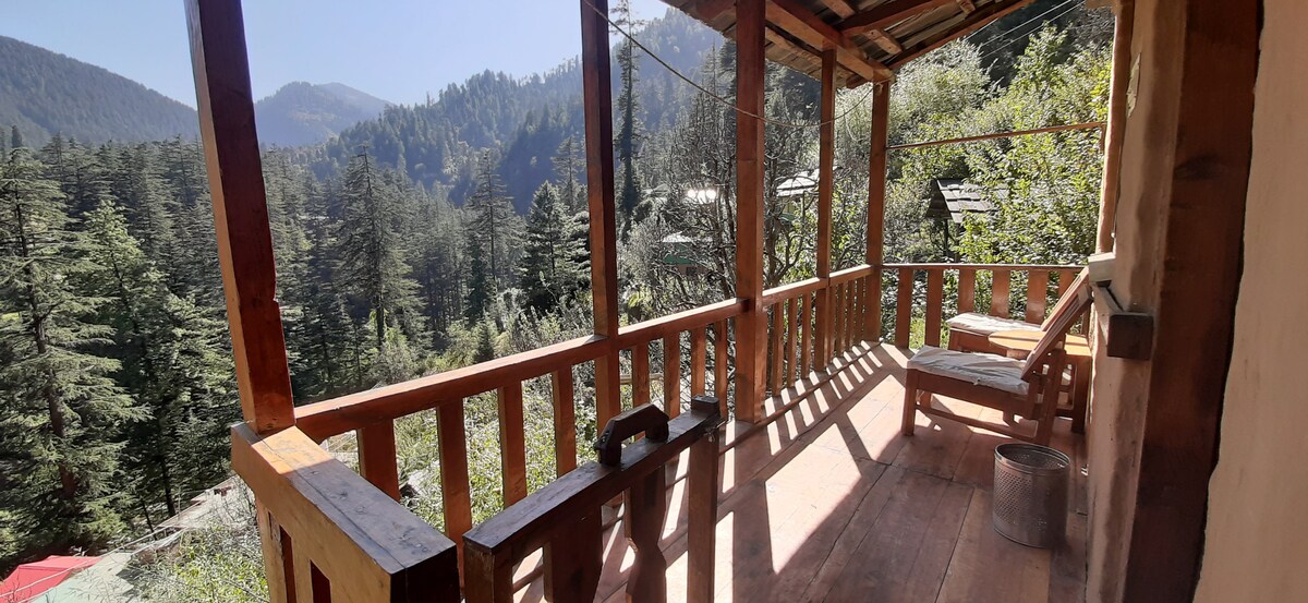 A wooden balcony extends from the cottage, offering unobstructed views of the tall cedar and pine trees surrounding the valley. Sunlight filters through the trees, illuminating the wooden seating area, which includes a bench and a table, enhancing the natural ambiance.