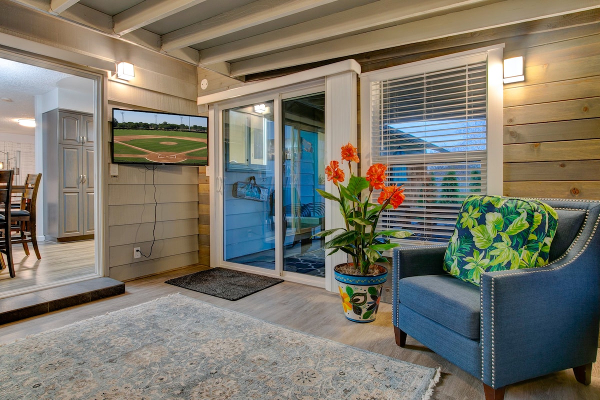 A sunroom features large sliding glass doors that provide natural light. A cozy blue armchair with tropical-patterned cushions is positioned beside a potted plant with vibrant orange flowers. A wall-mounted television displays a baseball game, enhancing the room's inviting ambiance.