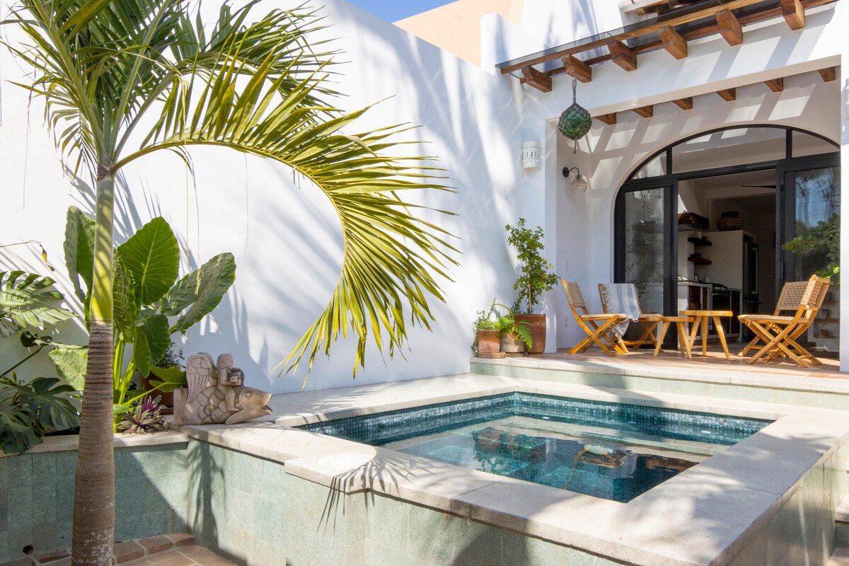 A serene outdoor space features a small pool surrounded by lush greenery and a palm tree. Wooden seating is arranged nearby under a shaded area, creating a cozy spot for relaxation and gathering. Sunlight filters through, enhancing the tranquil atmosphere.