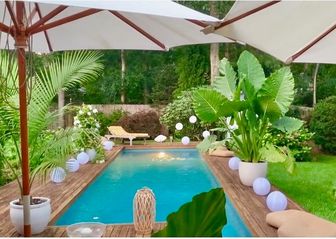 A heated lap pool is surrounded by lush greenery and oversized plants, creating a tranquil setting. Comfortable loungers are positioned on the wooden deck, complemented by decorative lanterns and soft lighting. Umbrellas provide shade, adding to the inviting atmosphere of the garden space.
