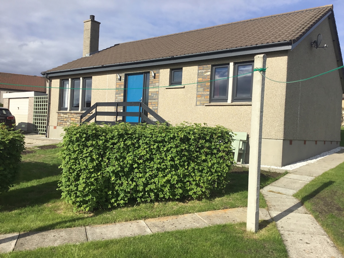 A single-story home features a light-colored exterior with stone accents. A bright blue door provides an inviting entrance. The landscaped front yard is bordered by low hedges, and a concrete walkway leads to the entrance. Windows, evenly spaced, allow natural light into the interior.
