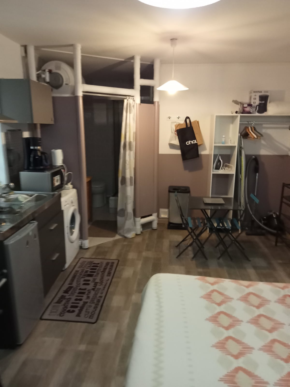 A compact living space is featured, showcasing a kitchenette with essential appliances and a dining table with two chairs. A washing machine is positioned nearby. The bathroom entrance is discreetly positioned, with light spilling into the room from a ceiling fixture.