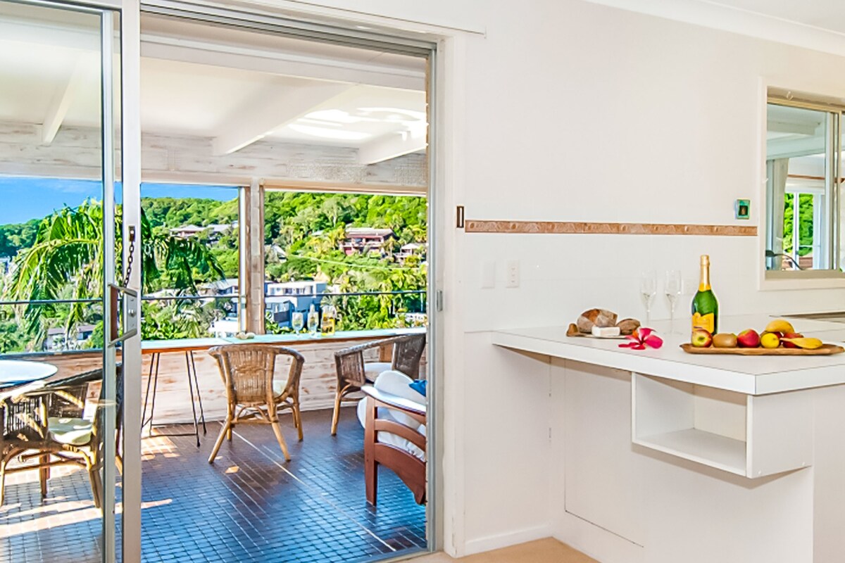 A bright kitchen space transitions to an outdoor dining area through sliding glass doors. A small breakfast bar is adorned with fresh fruit, bread, and champagne. Lush greenery is visible beyond the balcony, enhancing the airy feeling of the home.