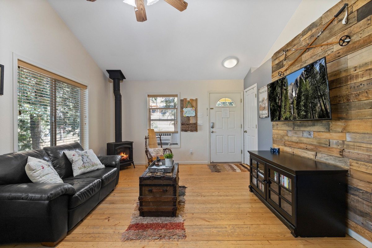 A spacious living area is furnished with a dark leather sofa and a rustic coffee table. Natural light floods in from large windows, while a cozy wood-burning stove is positioned in one corner. A flat-screen TV is mounted on a wooden accent wall, creating a warm ambiance.