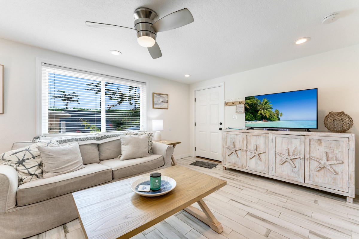 A bright living area contains a comfortable sofa adorned with coastal-themed pillows. A wooden coffee table sits in front, with a decorative tray placed on top. A television displays a serene beach scene, while light filters through the window with blinds.