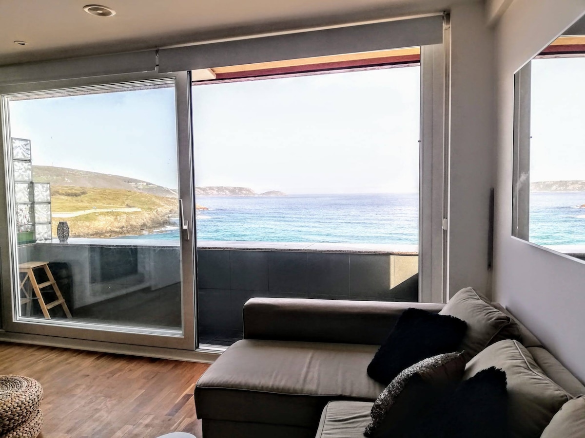 A living area with a comfortable sofa overlooks expansive views of the ocean through large glass sliding doors. Natural light fills the space, while coastal landscapes and rolling waves are visible in the distance, contributing to a serene environment.
